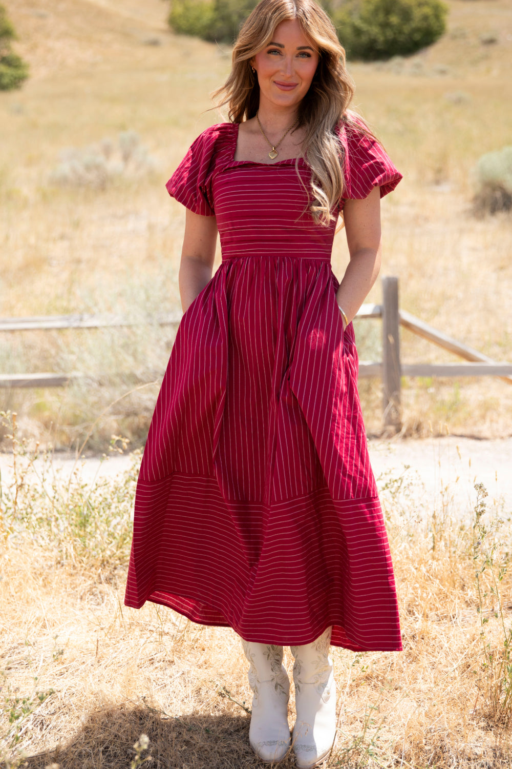 A woman stands outdoors in a sunny field wearing the Ivy City Co Julie Dress in Cranberry Pinstripe, featuring flutter sleeves, a square neckline, smocked bodice, and pockets; she pairs it with white cowboy boots.