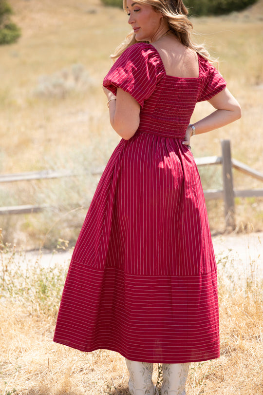 A woman stands outdoors on dry grass, wearing the Ivy City Co Julie Dress in Cranberry Pinstripe—a red, short-sleeve dress featuring stripes, a smocked bodice, and subtle puff sleeves. She faces away, looking over her shoulder, with a wooden fence and trees in the background.