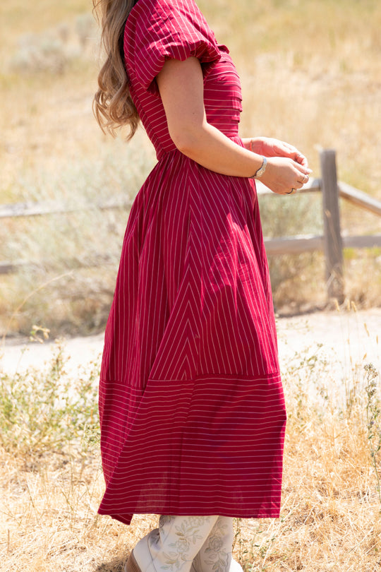A woman wearing the Ivy City Co Julie Dress in Cranberry Pinstripe, featuring puff sleeves and a smocked bodice, stands sideways outdoors in a dry grassy field with a wooden fence in the background. Only her body, not her face, is visible.