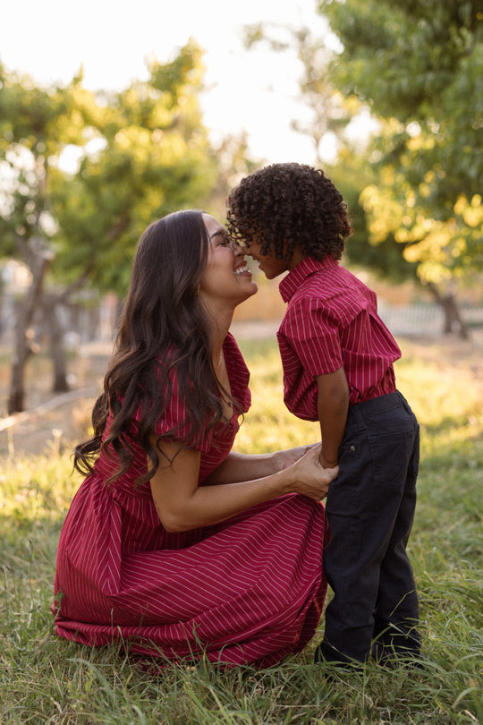 A woman kneels in the grass, smiling as she touches noses with a young boy. Both wear matching red outfits, with the woman in a charming Julie Dress in Cranberry Pinstripe by Ivy City Co, featuring a smocked bodice and puff sleeves. Trees and sunlight fill the background, suggesting a warm, joyful outdoor moment.
