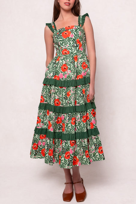 The model wears the Juno Dress in Green Floral, a sleeveless, tiered cotton dress with ruffled straps and bright red and white flowers. This ankle-length style is maternity friendly and styled with brown block-heeled shoes.