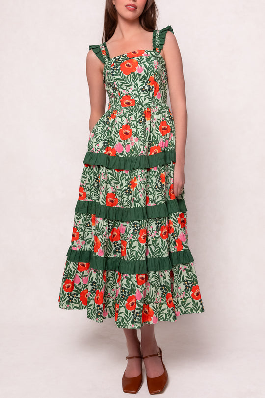 The model wears the Juno Dress in Green Floral, a sleeveless, tiered cotton dress with ruffled straps and bright red and white flowers. This ankle-length style is maternity friendly and styled with brown block-heeled shoes.