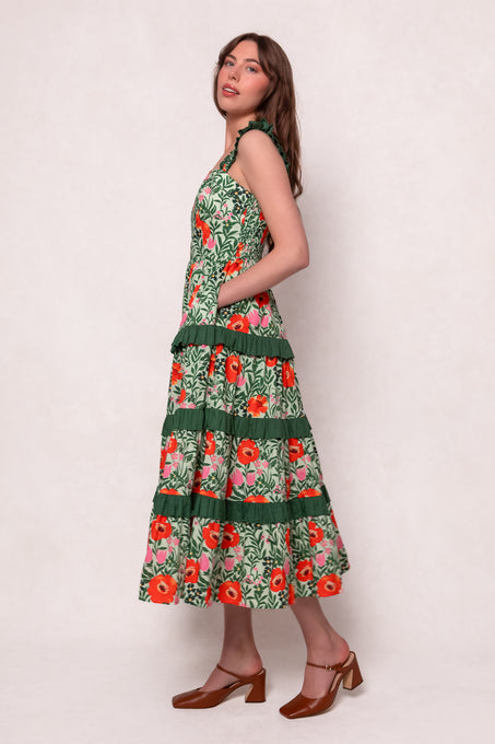 A woman stands sideways in the Juno Dress in Green Floral—a sleeveless cotton dress with bright red and orange flowers, ruffled tiers, and brown heeled shoes—against a plain light background.