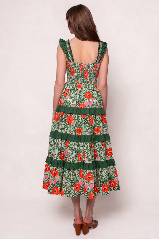 A woman with long brown hair, seen from the back, wears the Juno Dress in Green Floral—an ankle-length, sleeveless cotton dress with red floral patterns and tiered ruffles—paired with brown sandals against a plain light background.