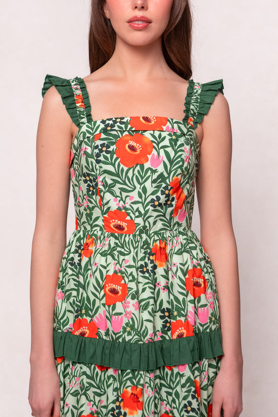 A woman wears the Juno Dress in Green Floral, a maternity-friendly sleeveless dress with a square neckline, ruffle straps, and a vibrant green floral print with orange and pink flowers, finished with a green ruffle waist trim.