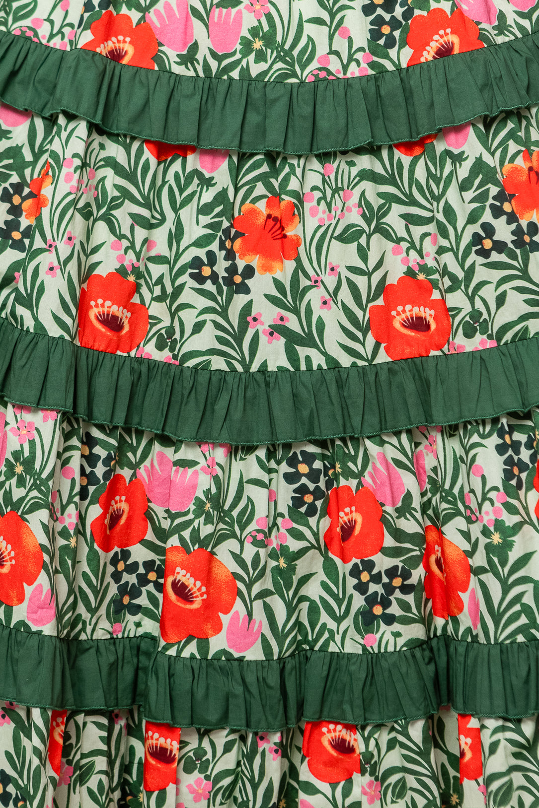 Close-up of the Juno Dress in Green Floral, featuring a colorful pattern of red, pink, and orange flowers with green leaves and horizontal dark green ruffles across soft cotton fabric.