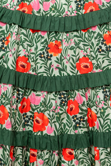 Close-up of the Juno Dress in Green Floral, featuring a colorful pattern of red, pink, and orange flowers with green leaves and horizontal dark green ruffles across soft cotton fabric.