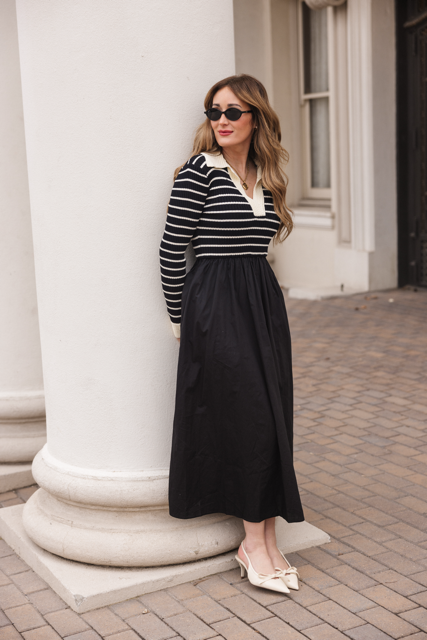 A woman in sunglasses leans against a white column, wearing the Adelina Dress in Black by Ivy City Co with cream heels, standing on patterned brick pavement near a building with large windows.