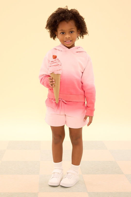 A young Black child with curly hair smiles while holding a large, handmade cardboard ice cream cone. They are wearing the Ivy City Co Mini Ash Hoodie in Pink Ombre and matching cotton fleece shorts, along with white socks and white sneakers, against a light yellow background.