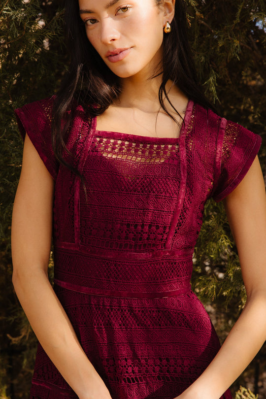 A woman with long dark hair wears the Ivy City Co Amory Dress in Burgundy, featuring a lace midi design and square neckline, paired with gold earrings as she stands in front of leafy green trees in natural sunlight.