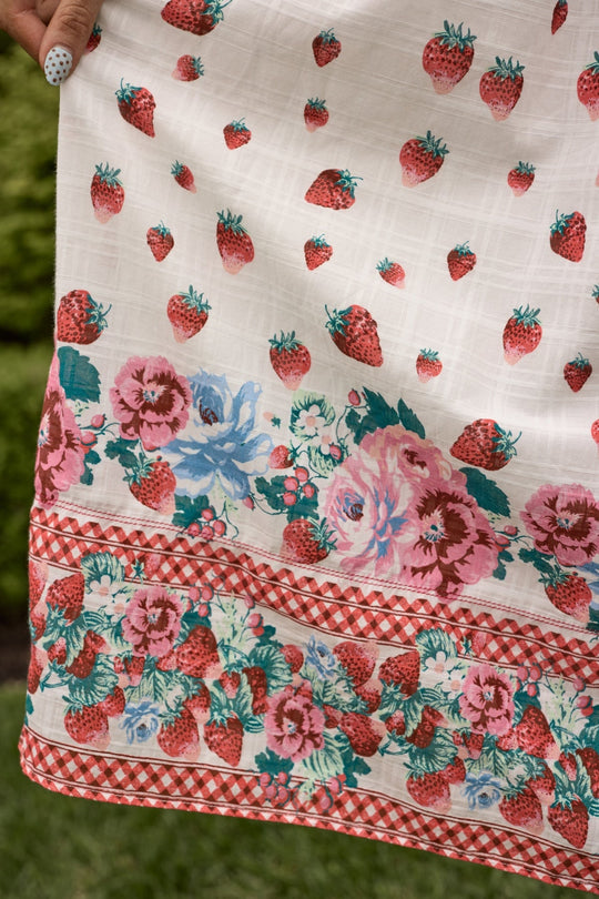 The Sadie Dress in Strawberry Vintage Tier by Ivy City Co features white cotton fabric adorned with red strawberries and pink, blue, and green floral patterns, finished with a charming red checkered trim. A manicured hand holds the fabric of this delightful strawberry print dress, while a backdrop of blurred green grass completes the scene.