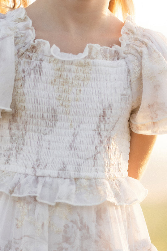 A close-up of a child wearing the Ivy City Co Mini Winnie Dress in Bright White Wispy Wildflower, showcasing its white ruffled design, puffed sleeves, textured smocked bodice, and delicate wildflower patterns—all beautifully illuminated by warm sunlight.