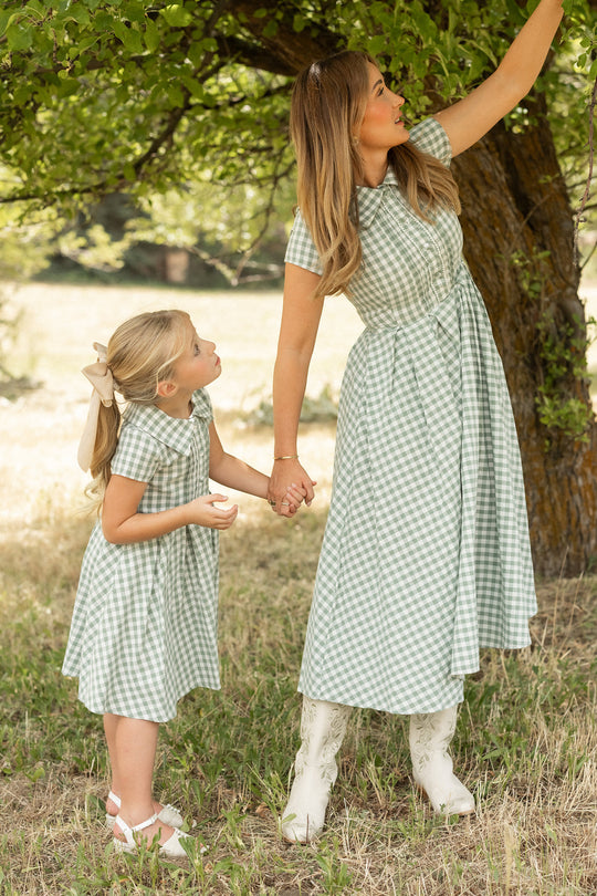 Wearing the Ivy City Co Mini Amber Dress in Sage Gingham, a woman and a young girl stand together under a tree on a sunny day. The woman, holding the girl’s hand, reaches up to touch the branches as the girl looks up at her.