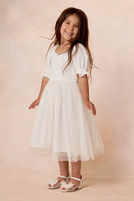 A smiling young girl with long brown hair wears the Ivy City Co Mini Ballerina Dress in White, featuring a flowing tulle skirt and paired with white sandals, standing against a soft beige background.