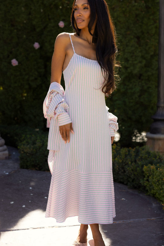 A woman stands outdoors among green bushes and pink flowers, wearing the Marbella Dress in Green & Pink Pinstripe by Ivy City Co— a sleeveless midi dress with thin straps and light pastel stripes — while holding a matching long-sleeve top.