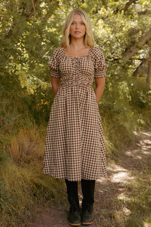 A woman stands on a sunlit dirt path in greenery, wearing the Kaitlyn Dress in Chocolate Gingham with puffed sleeves and black boots. This stylish look is also a maternity-friendly dress option.