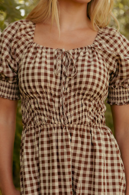 A woman in the Kaitlyn Dress in Chocolate Gingham, featuring a ruched waist and square neckline, stands outdoors against blurred greenery. This maternity-friendly dress blends comfort and effortless style. Her face is partially out of frame.