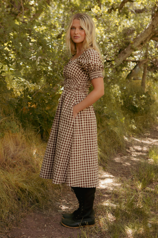 Wearing the Kaitlyn Dress in Chocolate Gingham and black boots, a woman stands on a dirt path among grass and trees, gazing calmly at the camera as sunlight streams through the leaves.