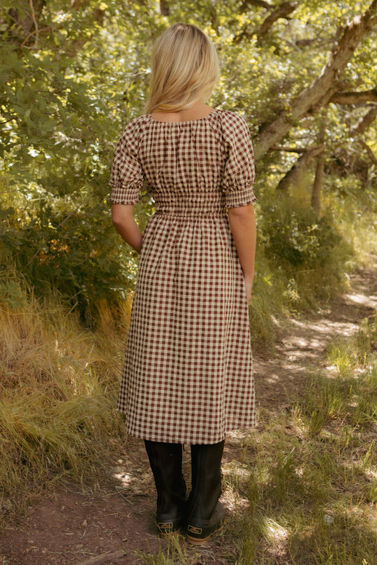 A person with blonde hair stands on a sun-dappled wooded path, wearing the Kaitlyn Dress in Chocolate Gingham with puff sleeves and black boots, facing away as sunlight filters through the trees.