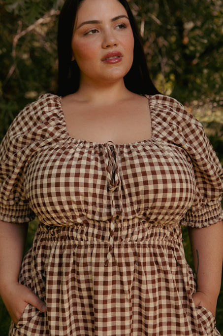 A woman with long dark hair wears the Kaitlyn Dress in Chocolate Gingham, featuring puffed sleeves and a square neckline, standing outdoors with greenery in the background.