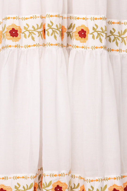 The Katerina Dress in White with Floral Trim by Ivy City Co features gathered, tiered cotton fabric accented with horizontal bands of red, yellow, and green embroidered floral designs.