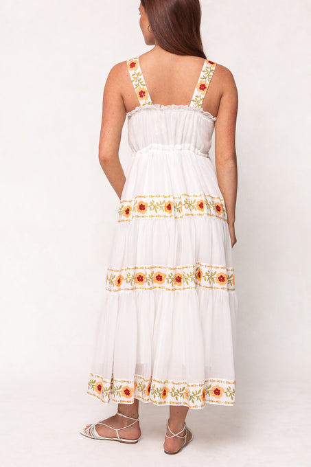 A woman with long brown hair, seen from the back, wears the Ivy City Co Katerina Dress in White with Floral Trim—a sleeveless, nursing-friendly dress with wide straps, a tiered skirt, and colorful floral embroidery—paired with white strappy sandals.
