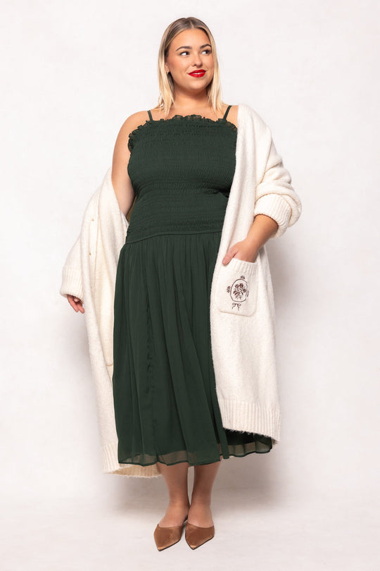 A woman stands against a white background wearing the Ivy City Co Kimberly Dress in Green, featuring a smocked bodice and midi length, paired with a white cardigan. She completes the look with tan pointed-toe flats. Her straight, shoulder-length blonde hair and red lipstick add to the stylish, maternity-friendly ensemble.