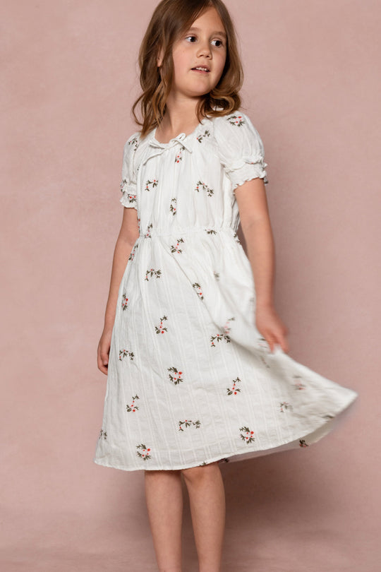 A young girl twirls gently against a light pink background, wearing the Ivy City Co Mini Jessie Dress in Ditsy Embroidery—featuring short sleeves and delicate small floral prints.
