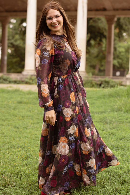 Wearing the Kelley Dress in Winter Floral by Ivy City Co, a woman stands smiling on the grass, turning slightly to the side. Tall columns and lush greenery appear softly blurred in the background.