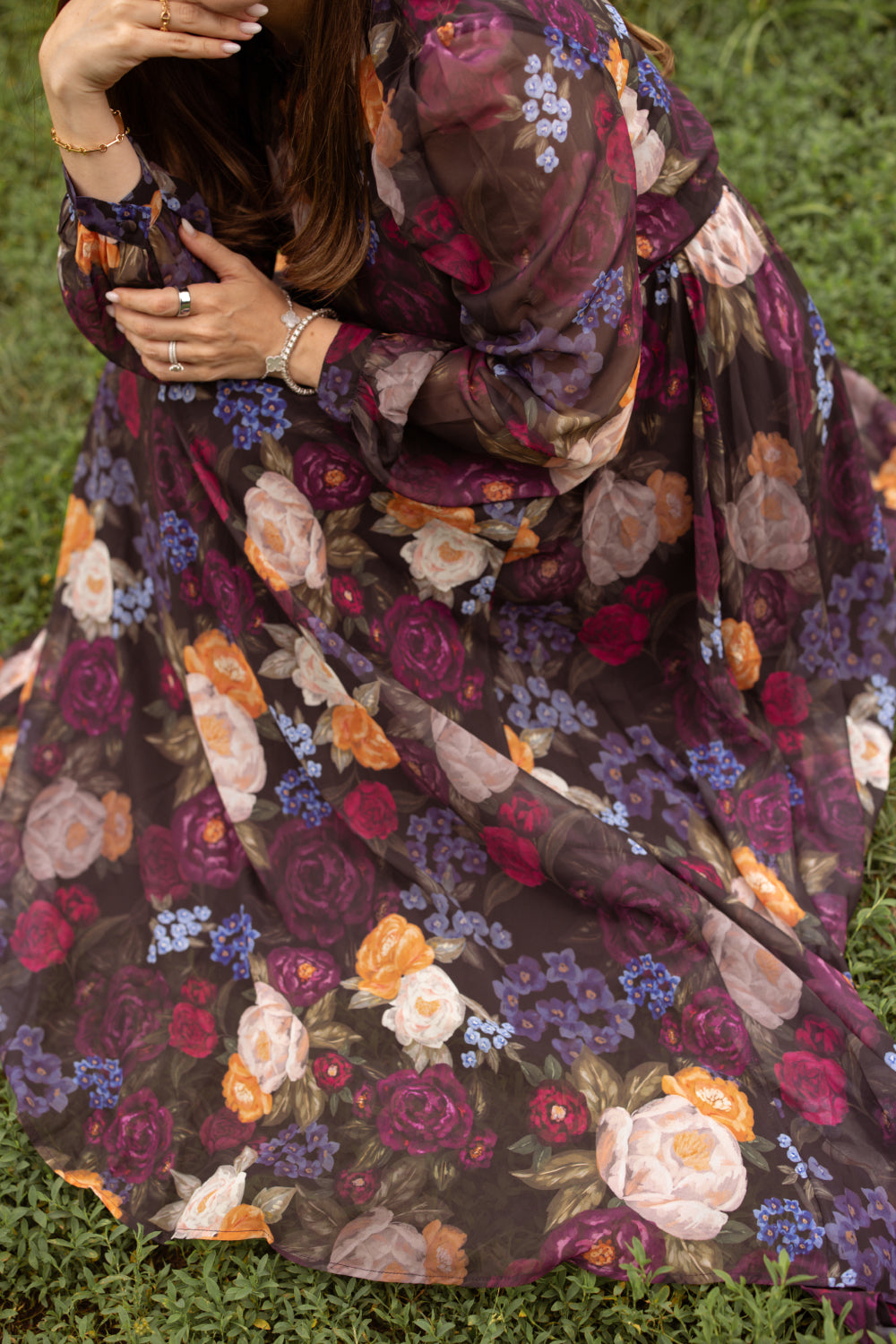 Wearing the Kelley Dress in Winter Floral by Ivy City Co—a long, flowing sheer purple maxi dress adorned with a colorful floral pattern—a woman sits gracefully on grass with her face partially out of frame, hands resting on her knee and jewelry on display, capturing the perfect moment for holiday portraits.