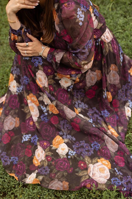 Wearing the Kelley Dress in Winter Floral by Ivy City Co—a long, flowing sheer purple maxi dress adorned with a colorful floral pattern—a woman sits gracefully on grass with her face partially out of frame, hands resting on her knee and jewelry on display, capturing the perfect moment for holiday portraits.