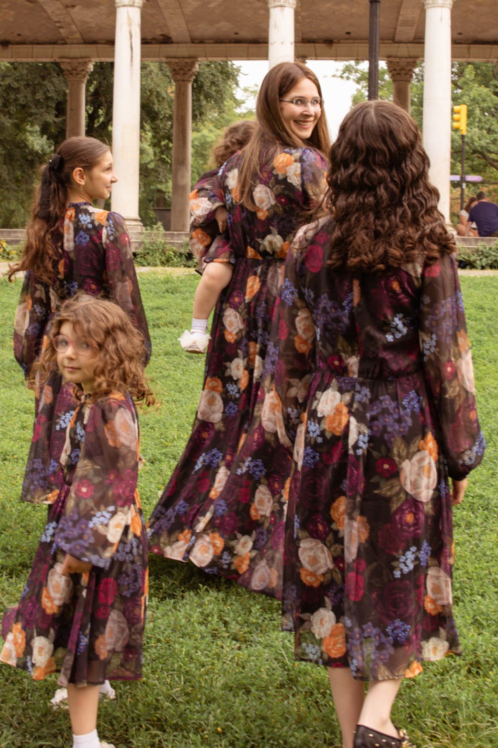 Five women stand on the grass in a park, all dressed in matching Kelley Dress in Winter Floral by Ivy City Co. Four face away while one turns back with a smile. With columns and lush greenery as the backdrop, it’s an ideal setting for holiday portraits.