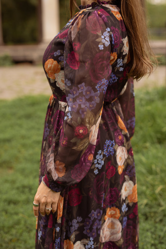 A woman wearing the Kelley Dress in Winter Floral by Ivy City Co stands outdoors on the grass. The image captures her from the shoulders down, showcasing the dress’s flowy fabric and vibrant winter floral pattern—an elegant option for holiday portraits.