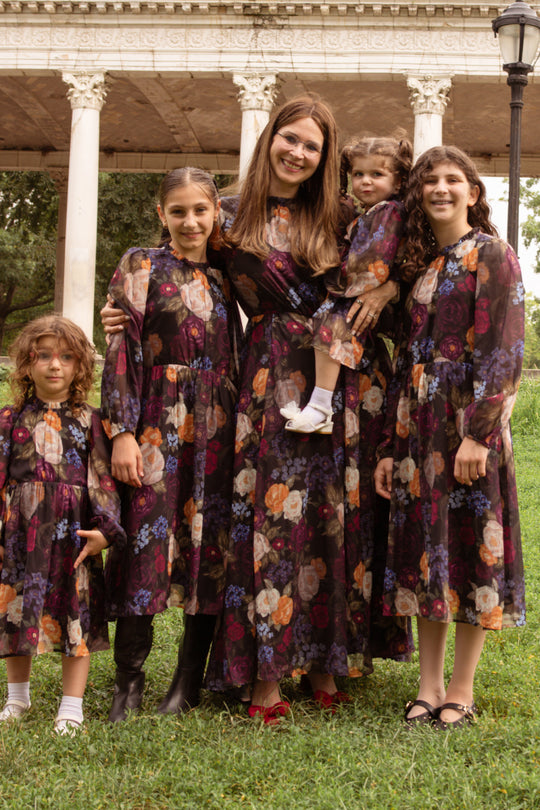 A woman and four girls stand on the grass, all wearing the Kelley Dress in Winter Floral by Ivy City Co. The woman holds a young child, and they are smiling in front of a white-columned structure—perfect attire for holiday portraits.
