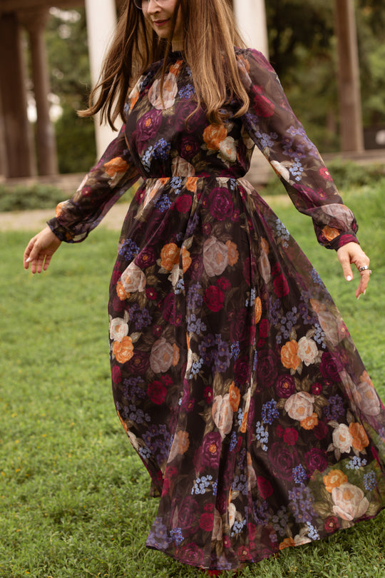 A woman with long brown hair twirls outdoors on green grass, wearing the Ivy City Co Kelley Dress in Winter Floral—a flowing, long-sleeved dress adorned with colorful patterns—set against a backdrop of trees and columns, perfect for capturing elegant holiday portraits.