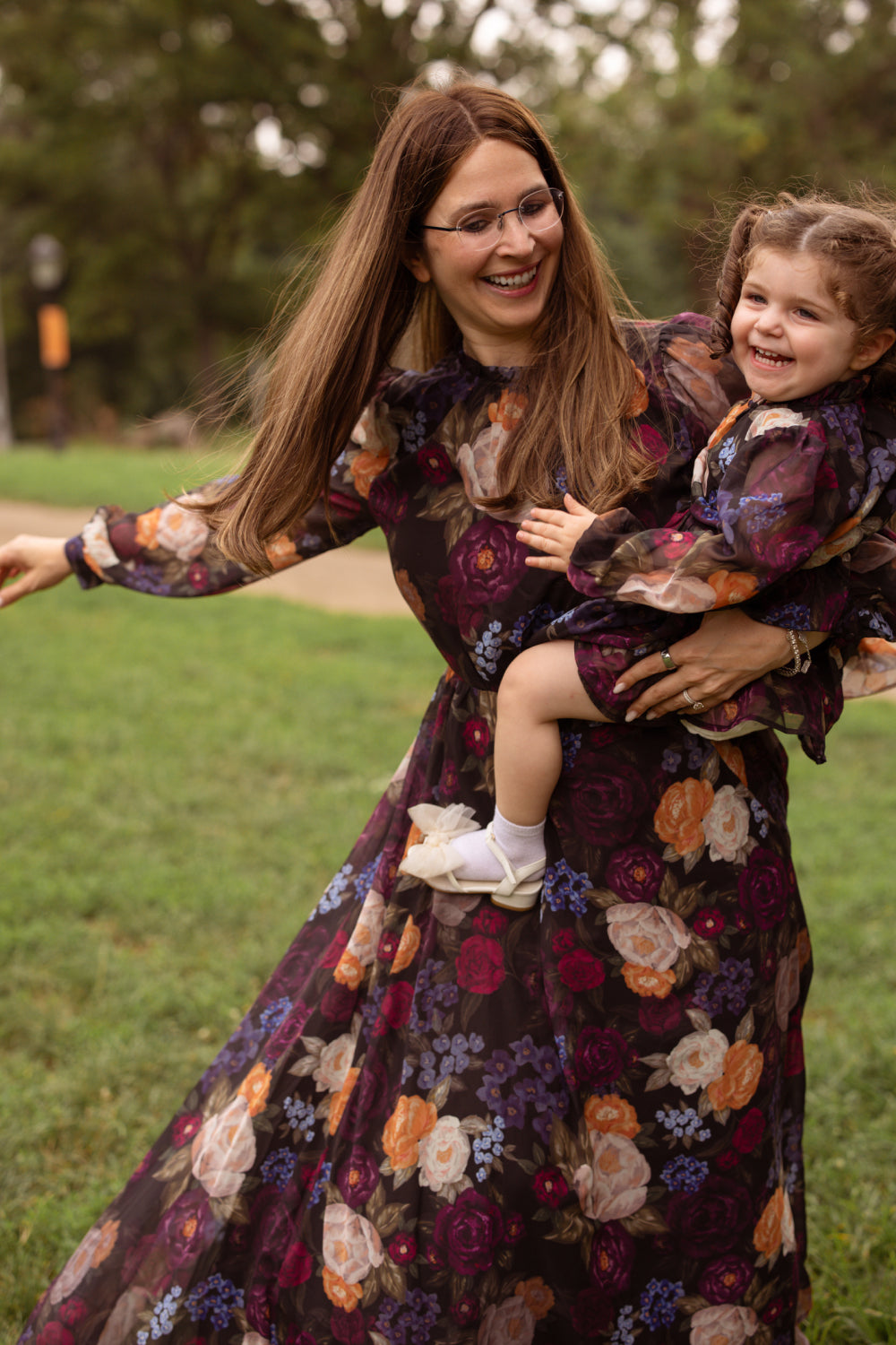 A woman and a young girl smile and spin together outside on the grass, both looking happy in matching Ivy City Co Kelley Dresses in Winter Floral.