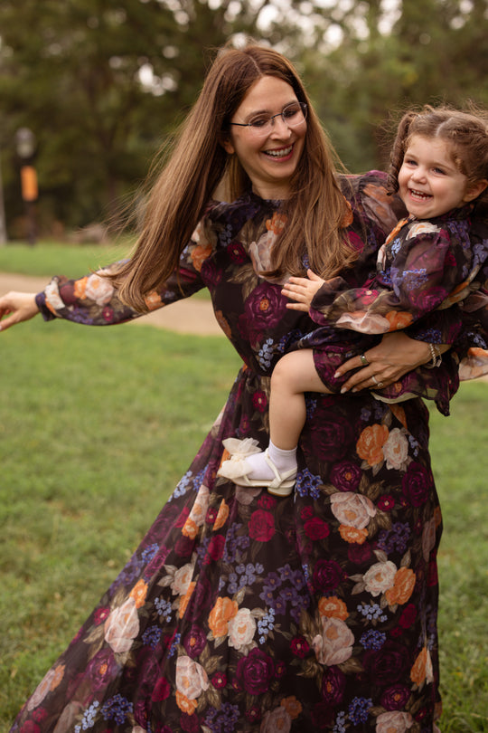 A woman and a young girl smile and spin together outside on the grass, both looking happy in matching Ivy City Co Kelley Dresses in Winter Floral.