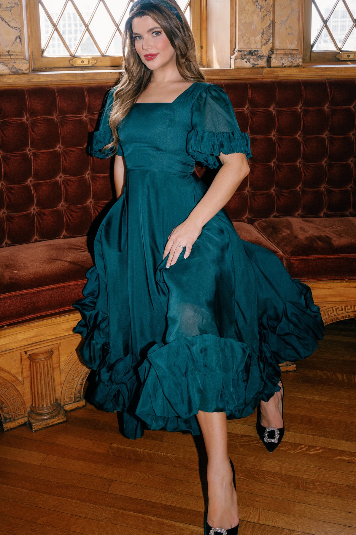 A woman poses indoors on a wooden floor by a tufted brown velvet bench and decorative windows, wearing the Ivy City Co Kendy Dress in Emerald paired with black heels.