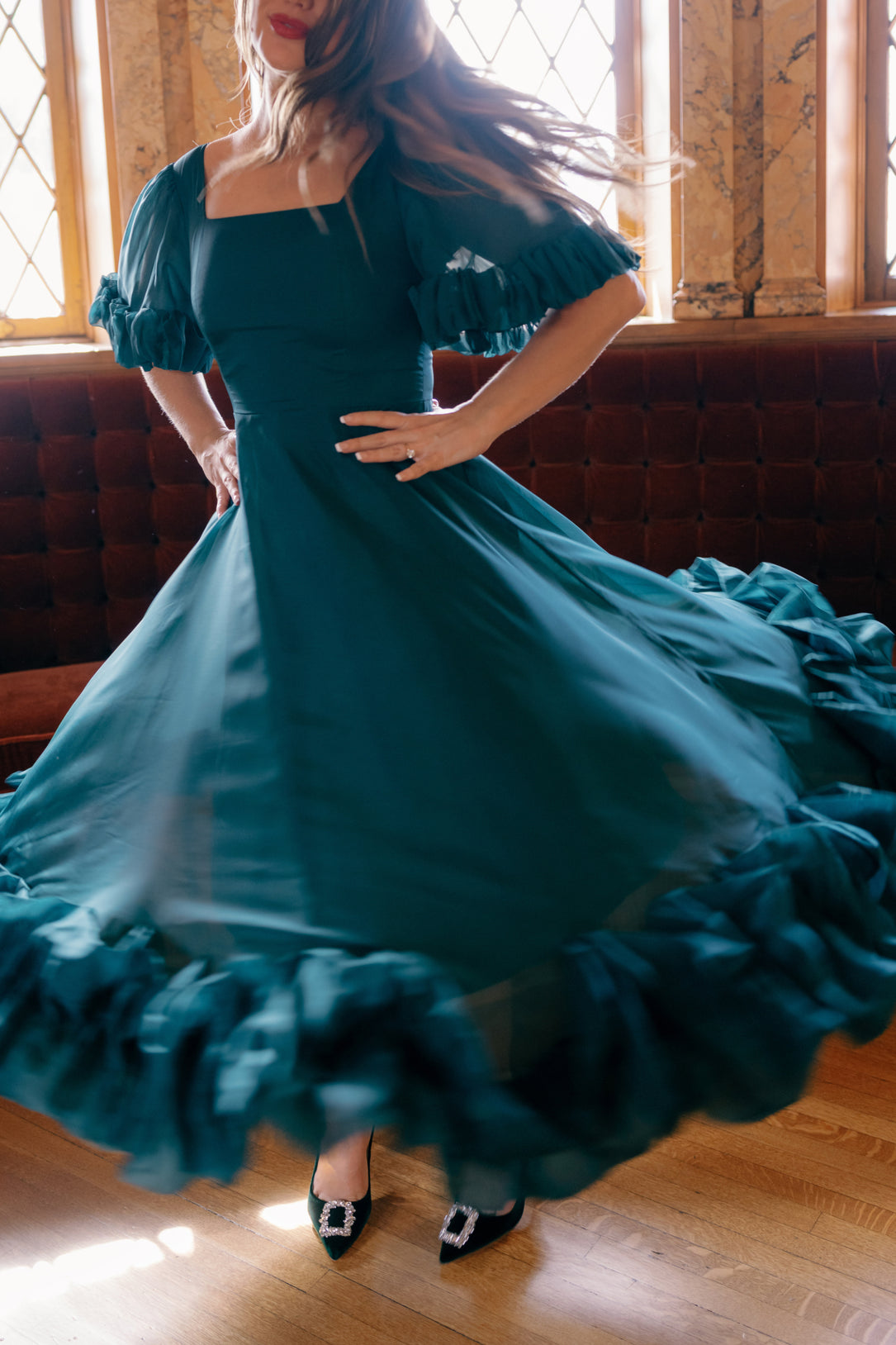 A woman twirls indoors in the Kendy Dress in Emerald by Ivy City Co—a flowing organza A-line dress with ruffled edges. Her hair and skirt ripple as she spins, black heels with jeweled buckles gleaming on her feet. She stands on a wooden floor near tall windows and a tufted couch.