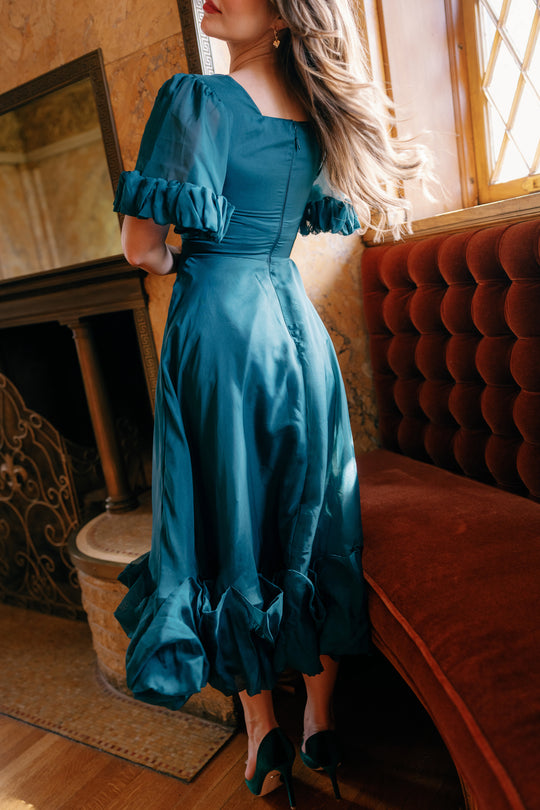 A woman in the Kendy Dress in Emerald by Ivy City Co—a vintage-style, organza A-line dress with ruffles—stands by a red velvet sofa and window, her wavy hair cascading down her back.