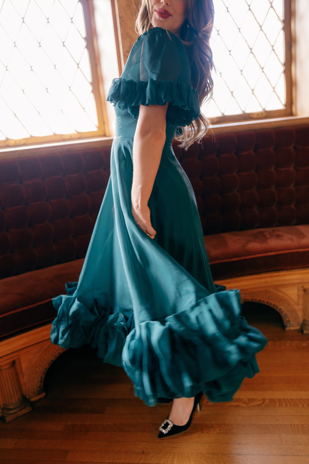 A woman twirls on a wooden floor by a tufted red velvet bench and large, diamond-paned windows, wearing the Ivy City Co Kendy Dress in Emerald.