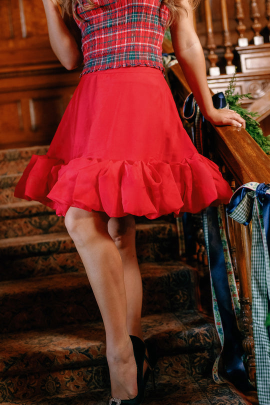 Wearing the Ivy City Co Kendy Skirt in Poinsettia and a red plaid top, a person stands on carpeted stairs holding the wooden banister, with decorated garland and ribbon adorning the railing. Face is not visible.