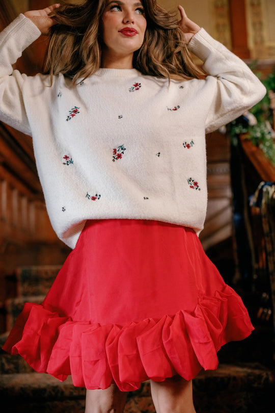 A woman wearing a white sweater with small floral designs and the Ivy City Co Kendy Skirt in Poinsettia, which features ruffles, stands indoors with her hands in her hair, gazing slightly off to the side.