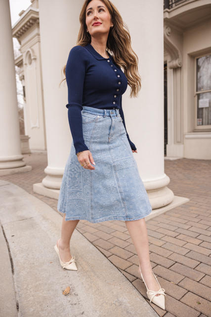 A woman with long hair walks outdoors near white columns, wearing a navy long-sleeve top, the Tarak Skirt in Light Wash Denim by Ivy City Co, and cream pointed shoes with bows.