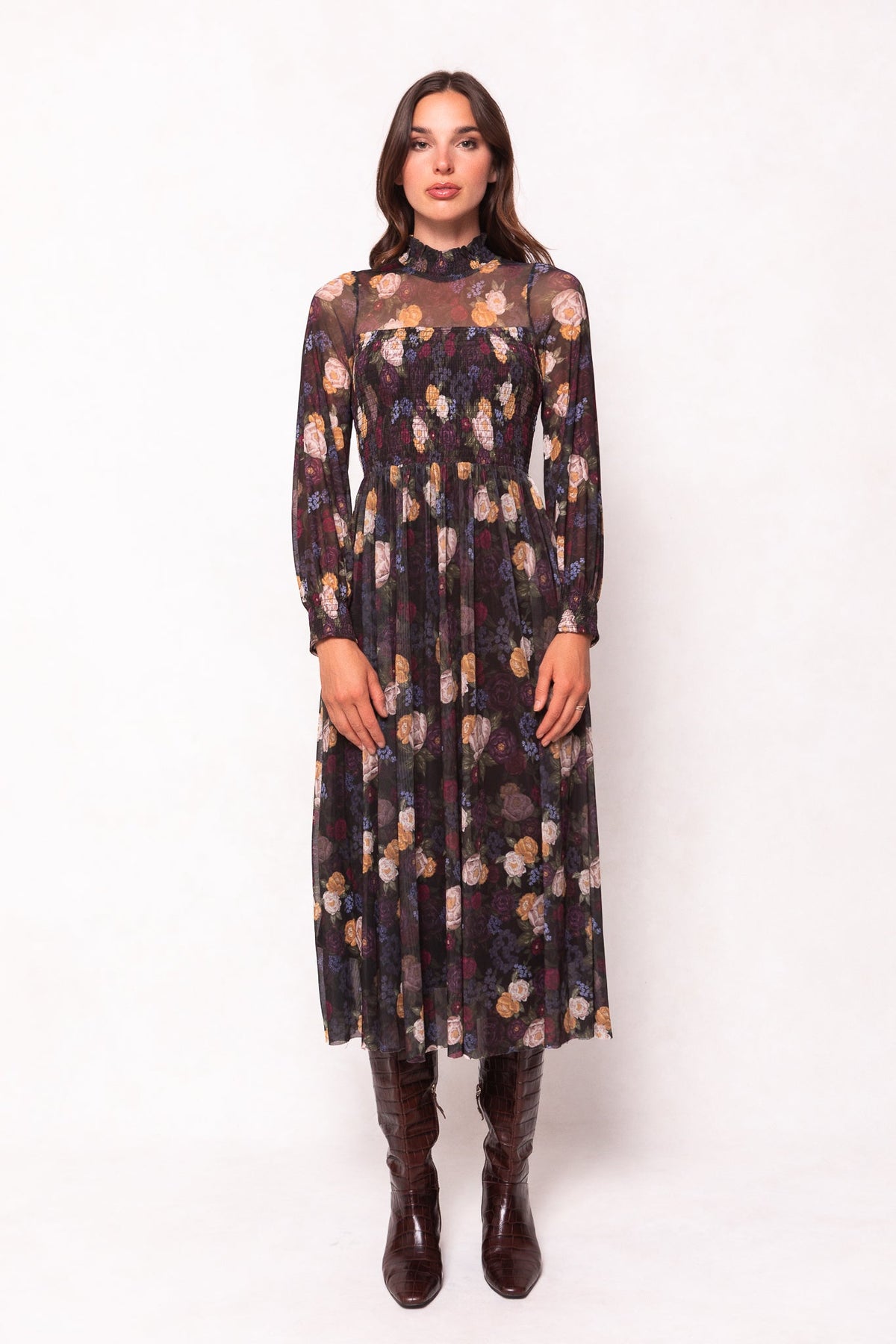 A woman stands against a plain light background, wearing the Ivy City Co Marla Dress in Winter Floral—a long-sleeved, ankle-length black dress with sheer detailing on the shoulders and chest. Perfectly maternity-friendly, she pairs it with brown knee-high boots.