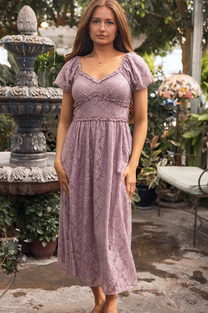 A woman with long brown hair wears the Ivy City Co Kiara Dress in Hyssop Lace and stands outdoors by a stone fountain, surrounded by plants and patio furniture.