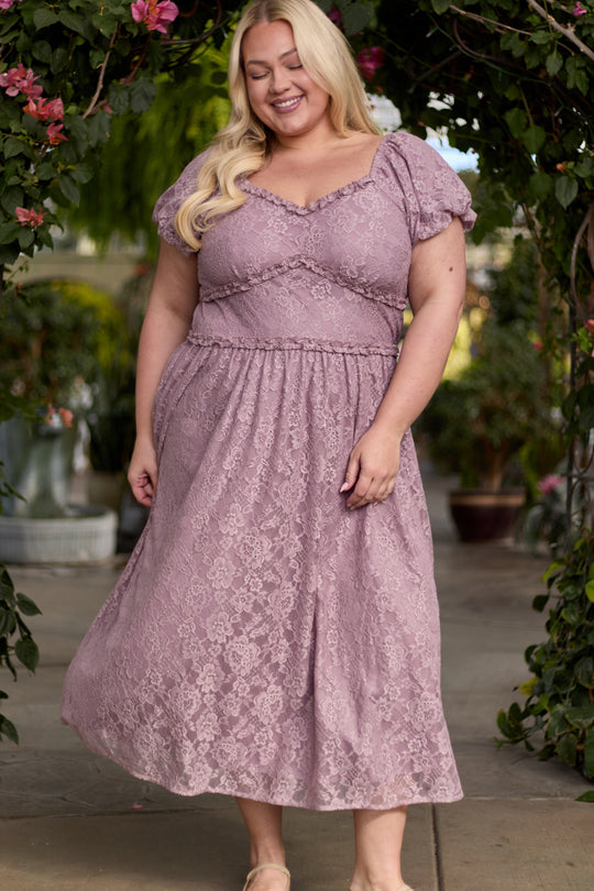 A smiling woman with long blonde hair wears the Ivy City Co Kiara Dress in Hyssop Lace, a dusty purple dress with short puff sleeves, standing outdoors beneath a floral archway adorned with pink blooms and greenery.