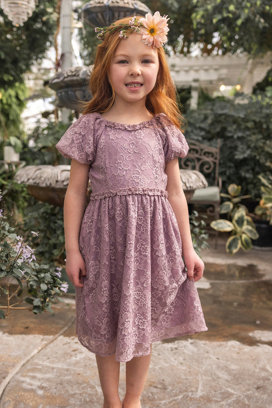 A young red-haired girl smiles at the camera outdoors, wearing the Ivy City Co Mini Kiara Dress in Hyssop Lace and a flower headband, surrounded by a lush garden.