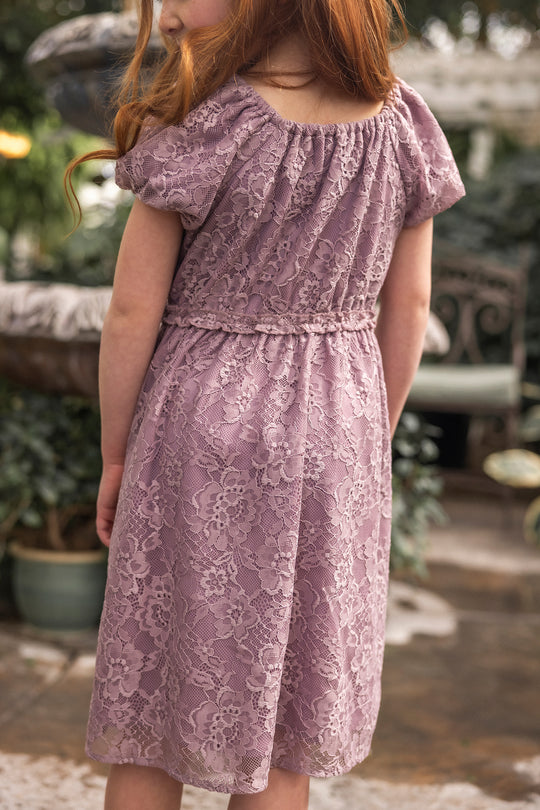 A young girl with long red hair stands outdoors among greenery and a stone fountain, wearing the Ivy City Co Mini Kiara Dress in Hyssop Lace—a charming choice for girls' parties.