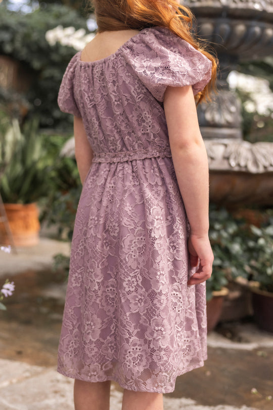 A woman with long red hair stands outdoors near a stone fountain, surrounded by greenery and potted plants. She is facing away, wearing the Ivy City Co Mini Kiara Dress in Hyssop Lace.